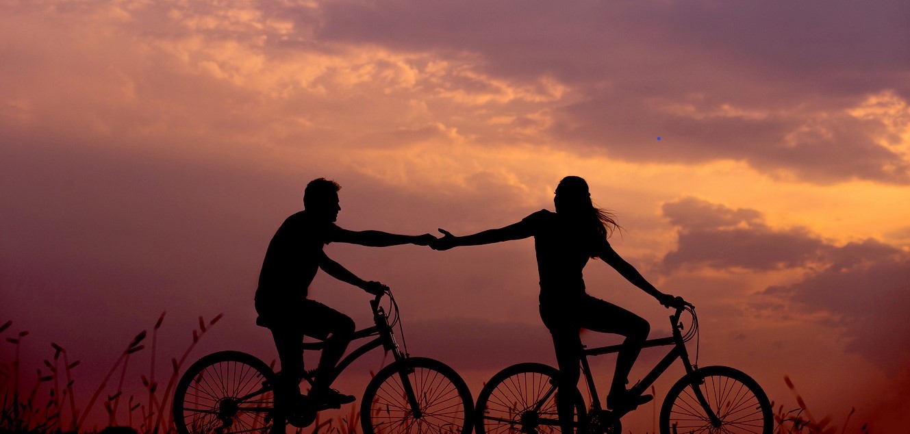 Two people reaching towards one another on bikes at sunset