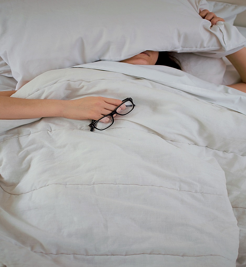 Person in bed with covers over this face holding glasses